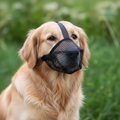 Dog wearing a black mesh muzzle outdoors with greenery in the background