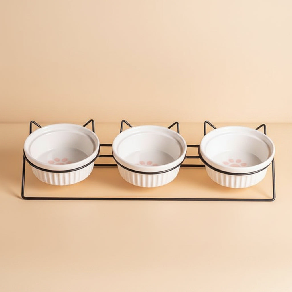 Three white ceramic pet bowls with paw prints on a black metal stand against an orange background