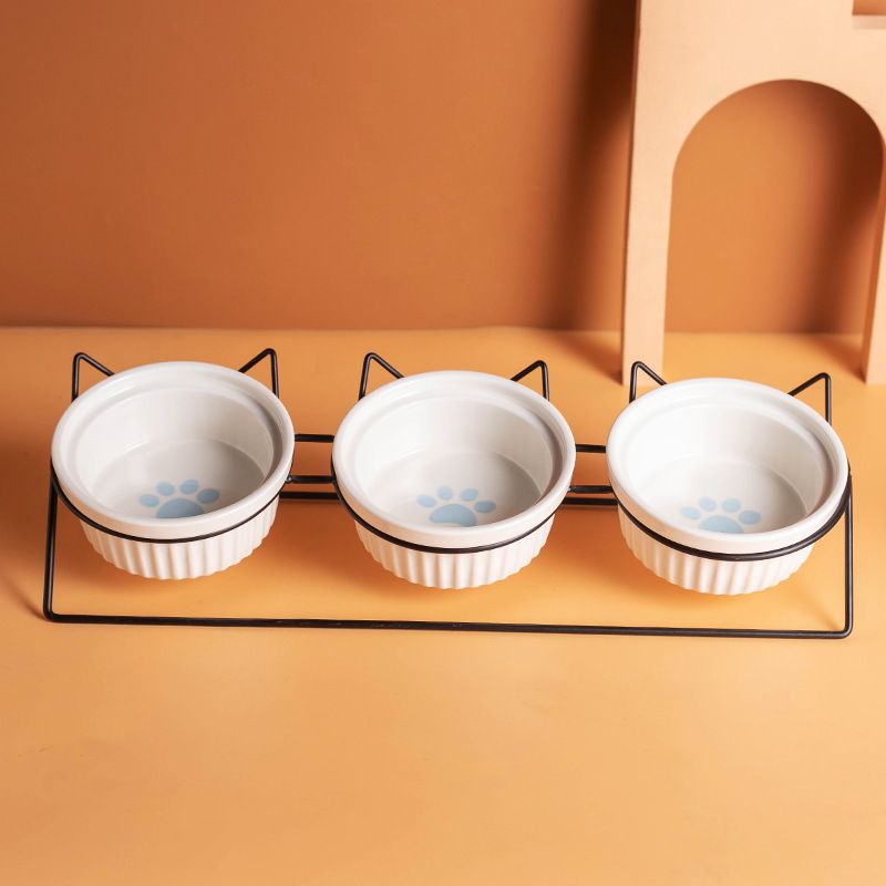 Three white pet bowls with paw prints on a black metal stand against an orange background