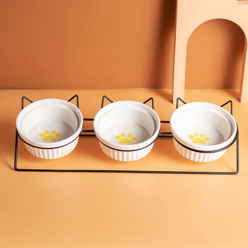 Three white pet bowls with yellow paw prints on a black metal stand against an orange background